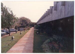 Calçadão lateral de edifício modernista, com fachada de pilares de concreto, jardim de arbustos à direita e árvores e carros estacionados à esquerda — P0015 — SESI — Ribeirão Preto