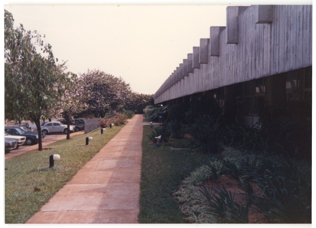 Calçadão lateral de edifício modernista, com fachada de pilares de concreto, jardim de arbustos à direita e árvores e carros estacionados à esquerda — P0015 — SESI — Ribeirão Preto — F009-P0015-1973-S03-D00041