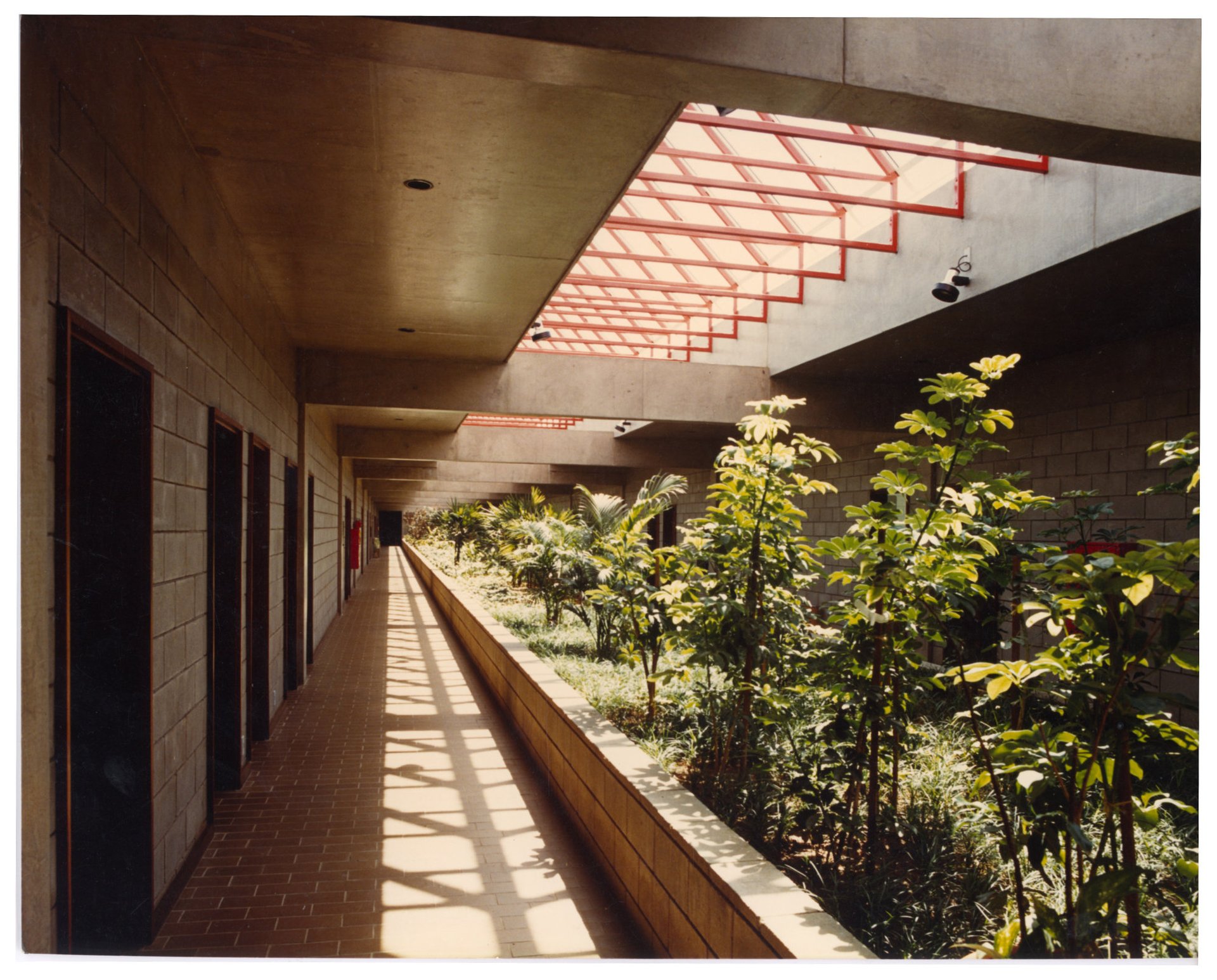 Corredor coberto com paredes de bloco de concreto, piso em tijolos, jardim interno lateral e estrutura metálica vermelha de clarabóia projetando sombras no piso — P0015 — SESI — Ribeirão Preto — F009-P0015-1973-S03-D00032