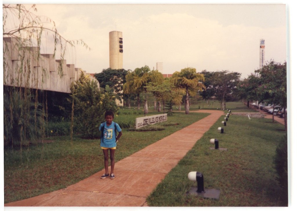 Criança posando em caminho de pedestres em área externa com edificação ao fundo e letreiro "SESI — P0015 — SESI — Ribeirão Preto — F009-P0015-1973-S03-D00034