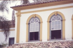Detalhe da fachada com janelas de arco pleno emolduradas em ocre e cinza, telhado de telhas cerâmicas em primeiro plano — P0024 — Fazenda Fernando Millan