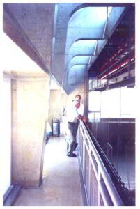 Duas pessoas observam o interior de um estádio a partir de um corredor com estrutura de concreto aparente e balaustrada metálica — P0131 — SESC - Piracicaba