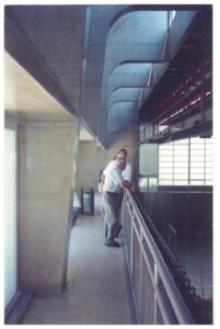 Duas pessoas observam o interior de um estádio a partir de um corredor com estrutura de concreto aparente e balaustrada metálica — P0131 — SESC - Piracicaba