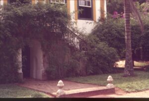 Fachada de edificação colonial com janelas emolduradas em amarelo, entrada em arco, caminho de pedras ladeado por remates ornamentais e vegetação exuberante — P0024 — Fazenda Fernando Millan