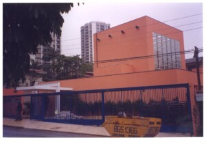 Fachada de edifício laranja com esquadrias de vidro, vista da rua, com grade metálica azul, caçamba de entulho amarela e prédios ao fundo — P0131 — SESC - Piracicaba