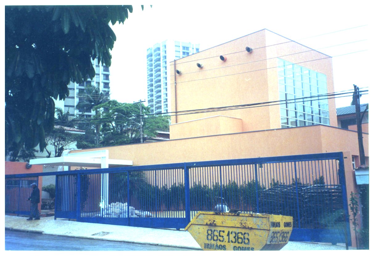 Fachada de edifício laranja com esquadrias de vidro, vista da rua, com grade metálica azul, caçamba de entulho amarela e prédios ao fundo — P0131 — SESC - Piracicaba — F009-P0131-0000-S03-D00037