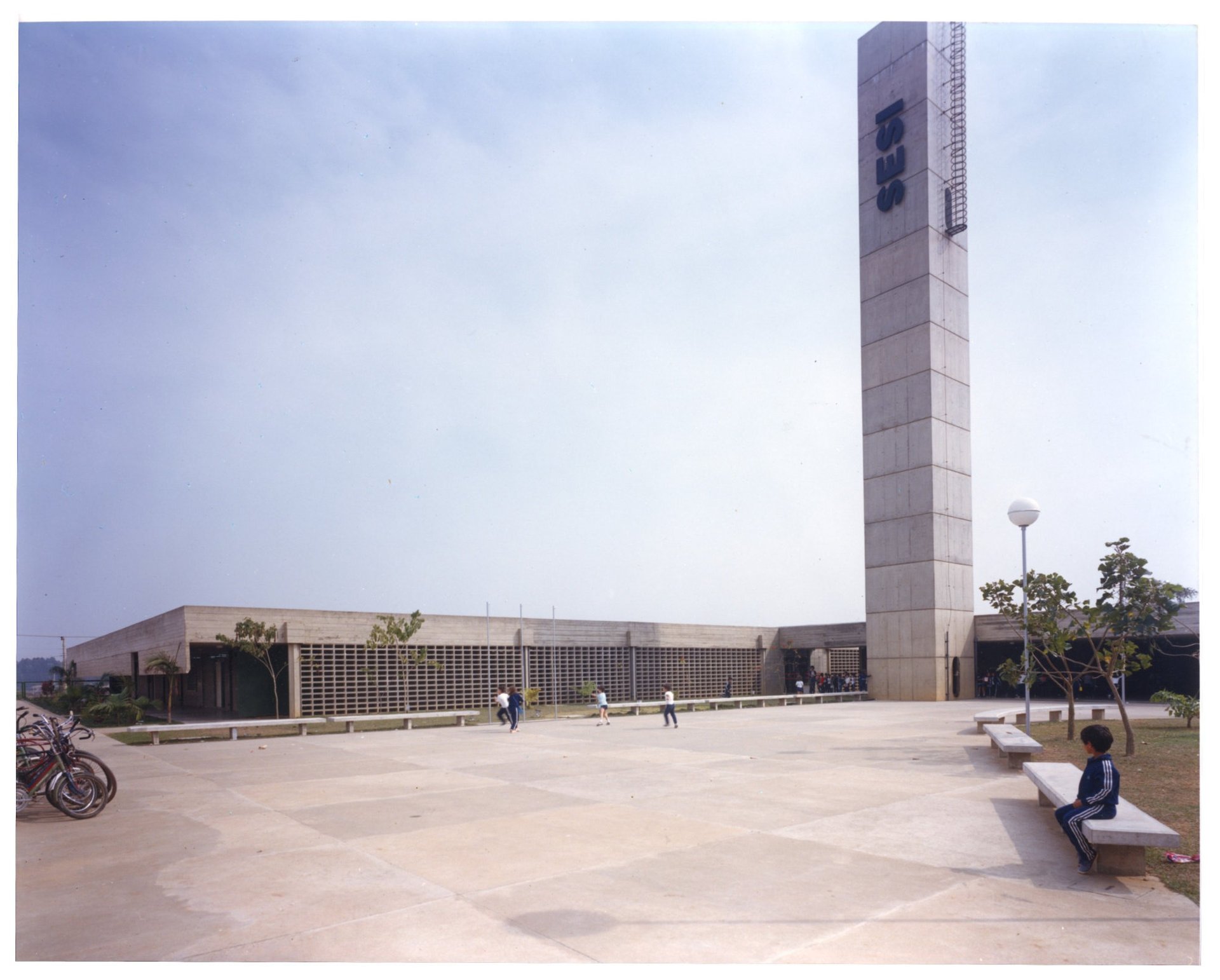 Fachada do edifício SESI, com pátio pavimentado, grades de ventilação e pessoas circulando nas proximidades — P0055 — SESI — São José dos Campos — F009-P0055-1979-S03-D00012