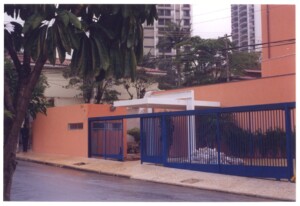 Fachada e portão de acesso de edificação em esquina, com muro na cor laranja e grade metálica azul, vista a partir da calçada — P0131 — SESC - Piracicaba