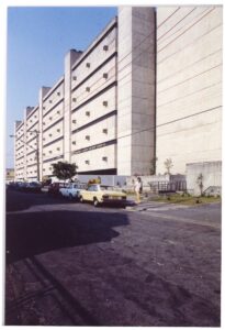 Fachada modulada em concreto aparente, vista em perspectiva ascendente, com veículos estacionados ao fundo e talude de terra ao fundo — P0018 — Correios — Sede Regional e Centro de Triagem