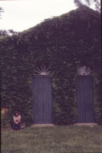 Fachada tomada por trepadeiras, com duas portas de madeira em arco com bandeiras em leque; pessoa sentada em primeiro plano sobre gramado — P0024 — Fazenda Fernando Millan