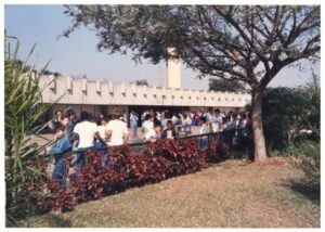 Grupo de pessoas reunidas junto a uma edificação de fachada ritmada por pilares, sob árvore de folhagem avermelhada — P0015 — SESI — Ribeirão Preto