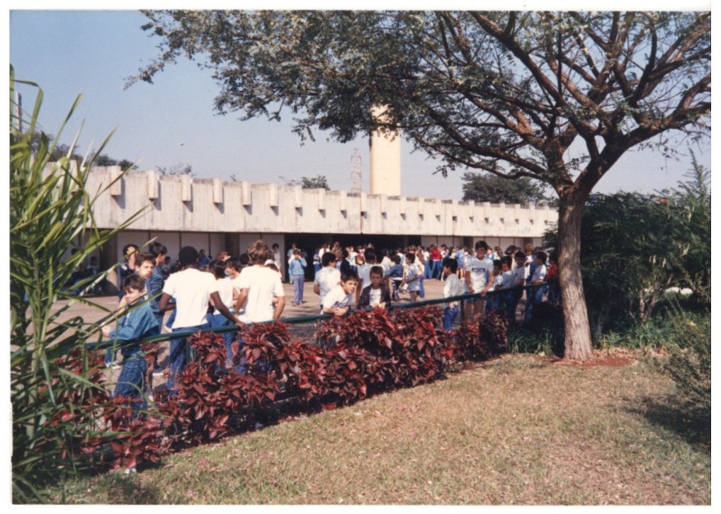 Grupo de pessoas reunidas junto a uma edificação de fachada ritmada por pilares, sob árvore de folhagem avermelhada — P0015 — SESI — Ribeirão Preto — F009-P0015-1973-S03-D00056