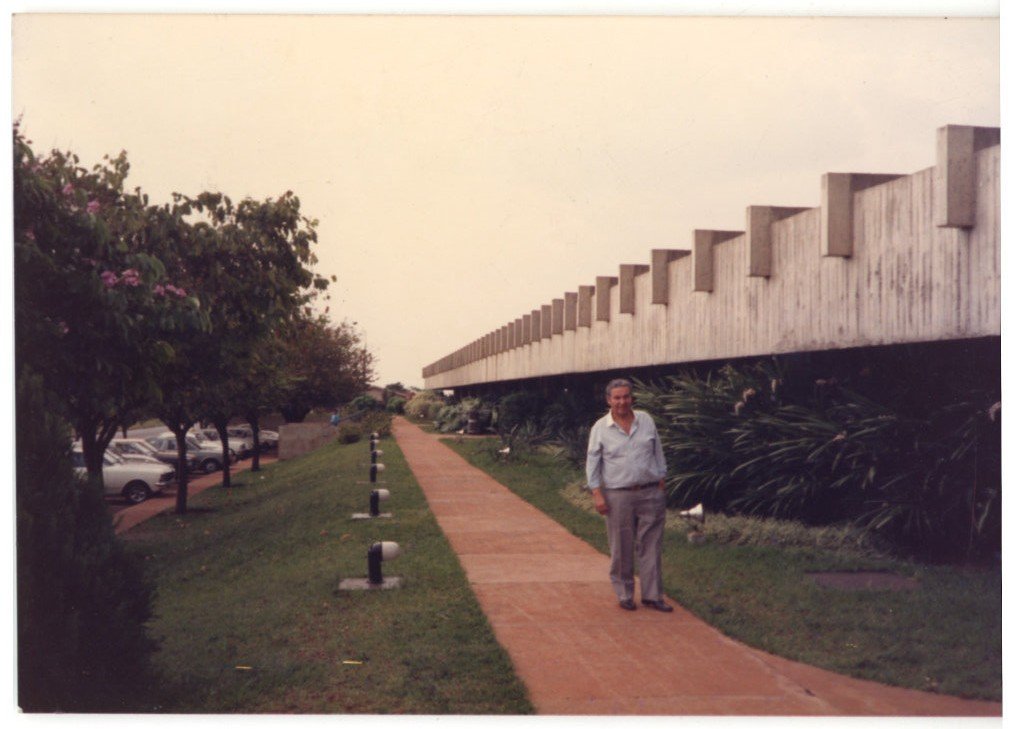 Homem posa em área externa ajardinada, próximo a uma estrutura de madeira, com muro de tijolos e árvores ao fundo — P0015 — SESI — Ribeirão Preto — F009-P0015-1973-S03-D00037