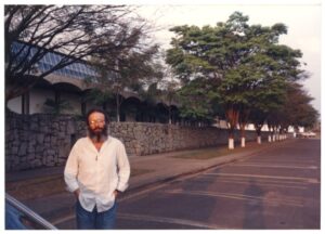 Cristiano Mascaro posa na calçada diante de muro de pedra e edifício com arcadas, em avenida arborizada — P0015 — SESI — Ribeirão Preto