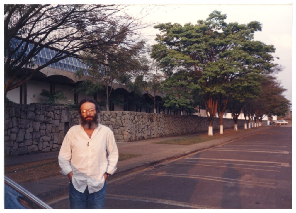 Cristiano Mascaro posa na calçada diante de muro de pedra e edifício com arcadas, em avenida arborizada — P0015 — SESI — Ribeirão Preto — F009-P0015-1973-S03-D00059