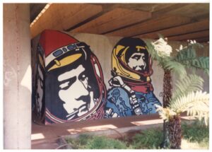 Mural de grafite com figuras de cosmonautas pintado em parede de concreto sob estrutura de viaduto, cercado por vegetação — P0015 — SESI — Ribeirão Preto