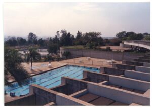 Piscina olímpica abrigada sob cobertura de concreto aparente, vista a partir da área externa com árvores ao redor — P0015 — SESI — Ribeirão Preto