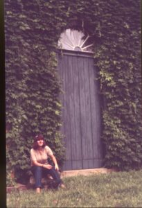 Porta de madeira com bandeira em leque, coberta por hera, com pessoa sentada em primeiro plano — P0024 — Fazenda Fernando Millan