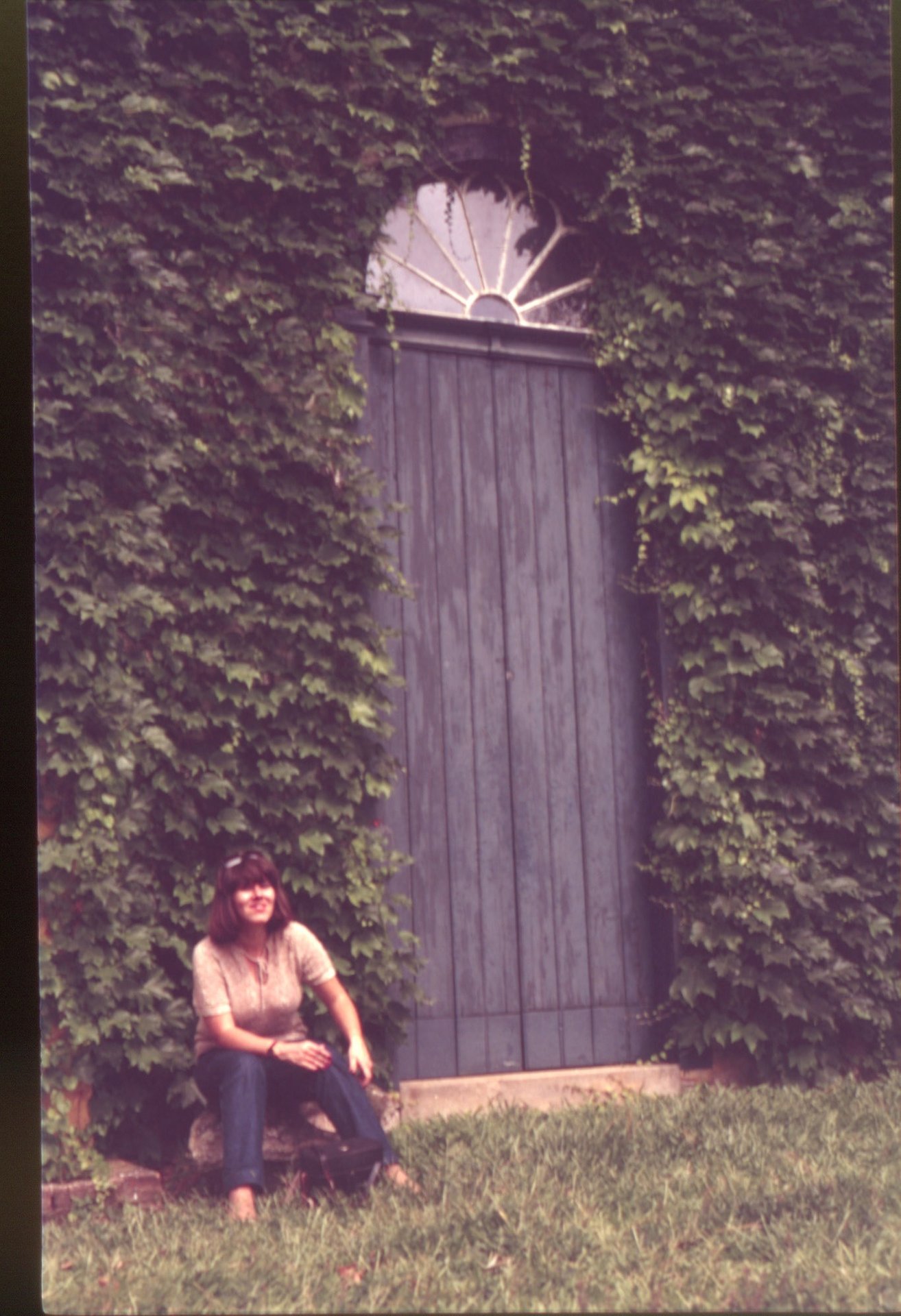 Porta de madeira com bandeira em leque, coberta por hera, com pessoa sentada em primeiro plano — P0024 — Fazenda Fernando Millan — F016-P0024-1981-S03-D00004