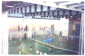 Quadra poliesportiva coberta com crianças jogando, vista de cima, estrutura metálica e redes de proteção ao redor da quadra — P0131 — SESC - Piracicaba