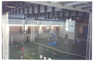 Quadra poliesportiva coberta com crianças jogando, vista de cima, estrutura metálica e redes de proteção ao redor da quadra — P0131 — SESC - Piracicaba
