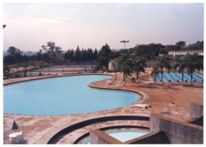 Vista aérea de conjunto de piscinas em clube ou complexo esportivo, com piscina principal de formato orgânico, piscina de raias, piscina circular e quadras ao fundo — P0015 — SESI — Ribeirão Preto