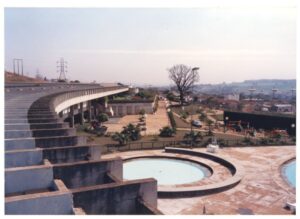 Vista aérea parcial de edifício com marquise curva em concreto, jardins, piscina circular e área de lazer, com a cidade ao fundo — P0015 — SESI — Ribeirão Preto