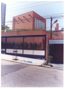 Vista da rua do portão e fachada superior de edifício em tom terracota, com muro, vegetação e poste de iluminação — P0131 — SESC - Piracicaba