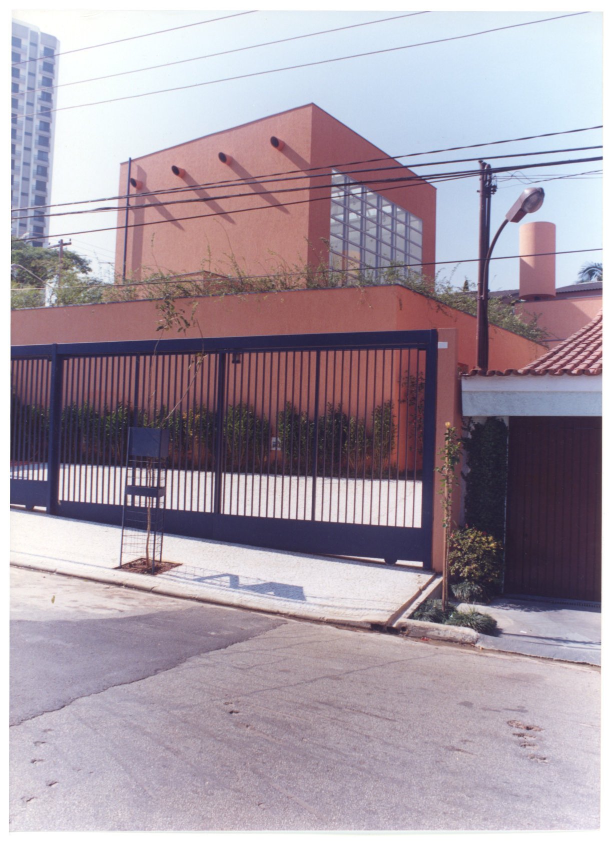 Vista da rua do portão e fachada superior de edifício em tom terracota, com muro, vegetação e poste de iluminação — P0131 — SESC - Piracicaba — F009-P0131-0000-S03-D00011