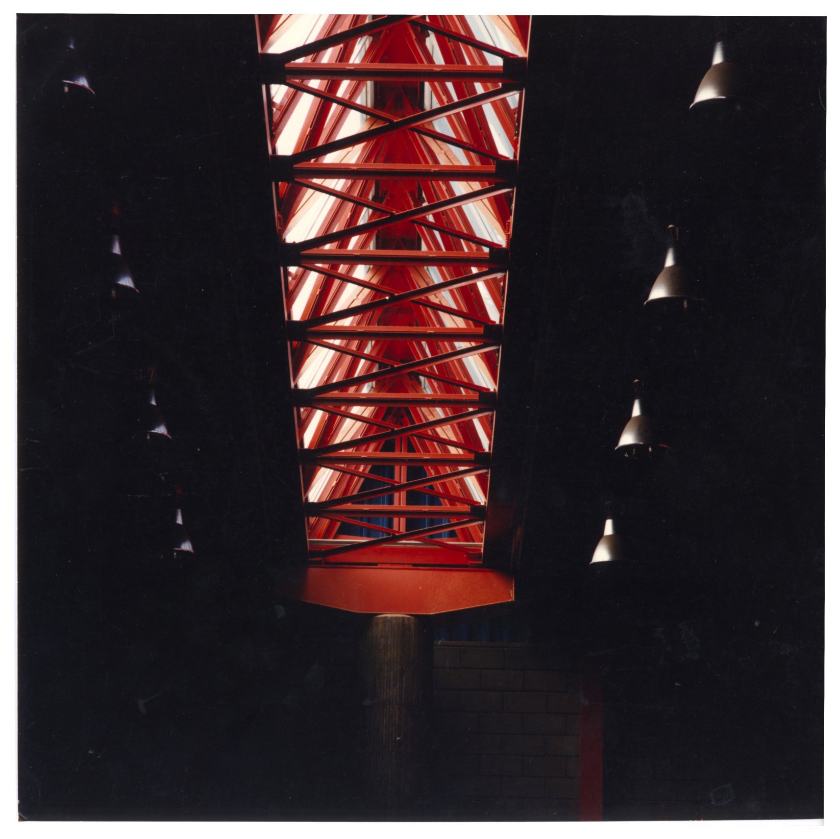 Vista de baixo para cima de clarabóia estruturada em treliças metálicas vermelhas, com luminárias suspensas em primeiro plano, ambiente interno escuro — P0060 — SESI — Marília — F009-P0060-1984-S03-D00031