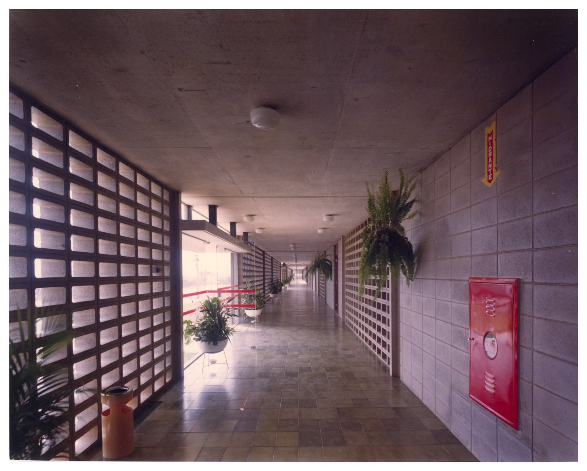 Vista de corredor interno com piso em grelha, plantas suspensas e placa de extintor/sinalização de rota de fuga — P0055 — SESI — São José dos Campos — F009-P0055-1979-S03-D00016