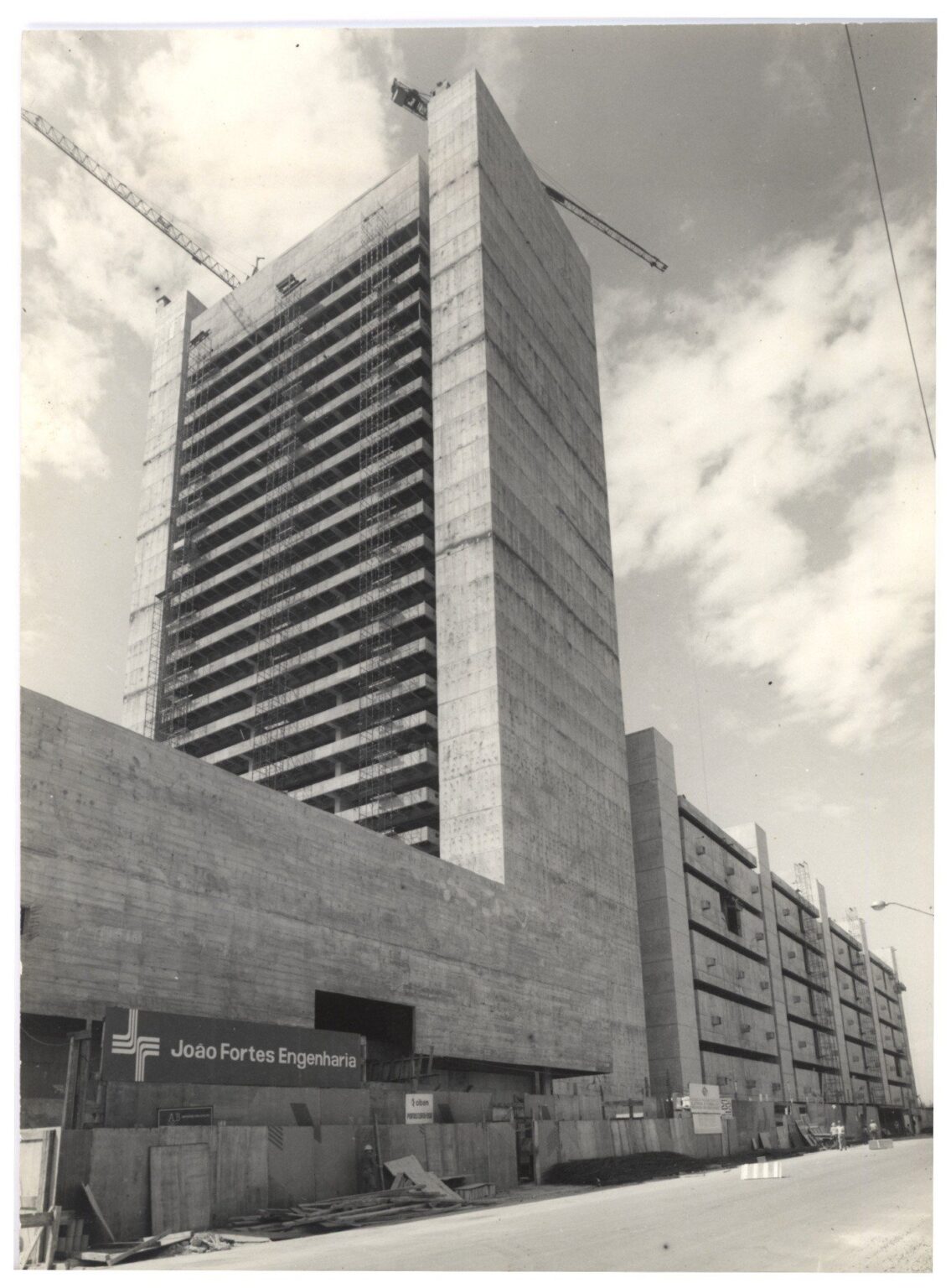 Vista de edifício em construção, com estrutura de concreto aparente e placa da empreiteira "João Fortes Engenharia — P0018 — Correios — Sede Regional e Centro de Triagem — F009-P0018-1975-S03-D00002