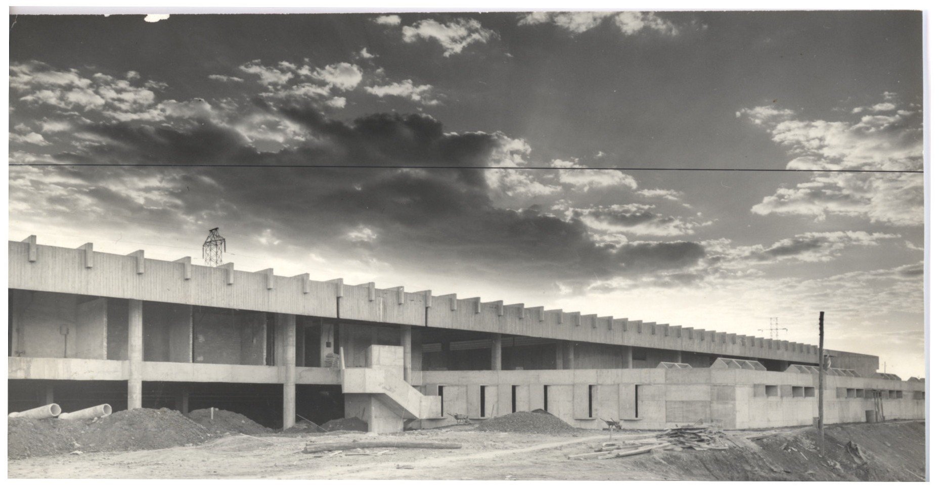 Vista de edifício em obras, com estrutura de concreto aparente, pilares e vigas, terreno ao fundo e céu com nuvens (imagem invertida) — P0015 — SESI — Ribeirão Preto — F009-P0015-1973-S03-D00020