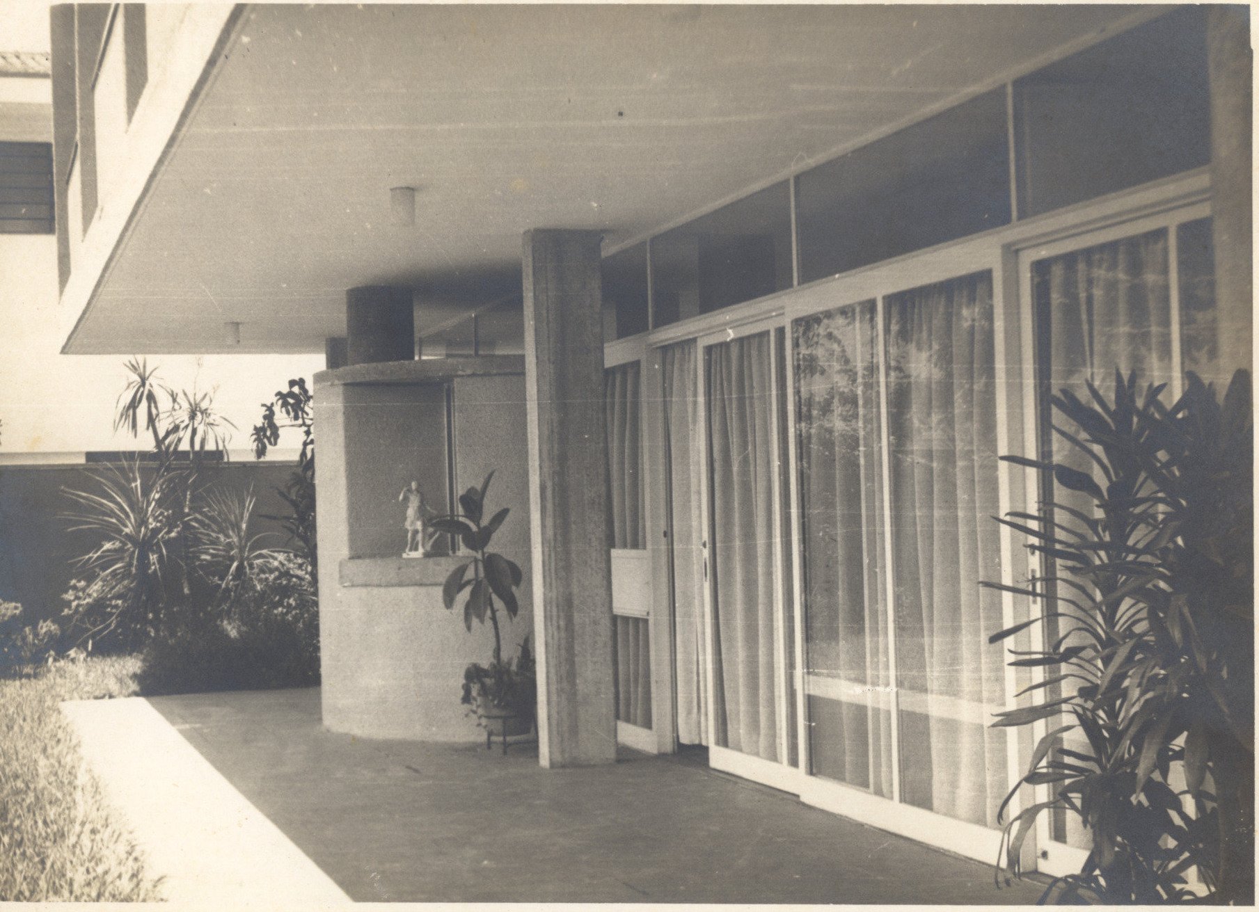 Vista de fachada envidraçada de edifício, com vegetação ao fundo e em primeiro plano — P0001 — Residência Aecio Aeouche de Toledo — F004-P0001-1960-S03-D00003