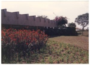 Vista de jardim com muro de concreto em arcos, canteiro de flores vermelhas e árvores ao fundo — P0015 — SESI — Ribeirão Preto