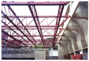 Vista de obra em andamento: estrutura metálica treliçada pintada de vermelho junto a elemento estrutural de concreto em forma de arco, com vegetação e edifícios ao fundo — P0131 — SESC - Piracicaba