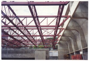 Vista de obra em andamento: estrutura metálica treliçada pintada de vermelho junto a elemento estrutural de concreto em forma de arco, com vegetação e edifícios ao fundo — P0131 — SESC - Piracicaba