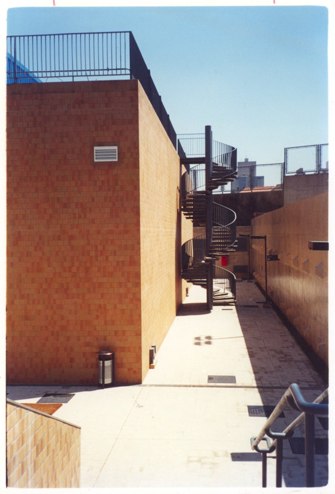 Vista de passarela coberta por estrutura metálica ondulada, piso cerâmico e parede revestida em cerâmica, ao lado de muro claro — P0131 — SESC - Piracicaba — F009-P0131-0000-S03-D00003