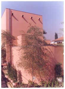 Vista de pátio e fachada de edificação térrea em tom terracota, com árvores, vasos de plantas e muro ao fundo — P0131 — SESC - Piracicaba