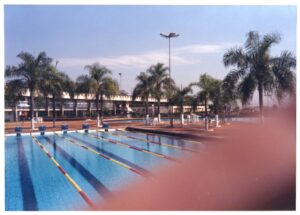 Vista de piscina de raias com edifício e palmeiras ao fundo, sob céu claro — P0015 — SESI — Ribeirão Preto