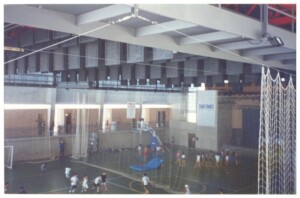 Vista de quadra poliesportiva coberta com estrutura metálica aparente, redes de proteção e pessoas praticando esportes — P0131 — SESC - Piracicaba
