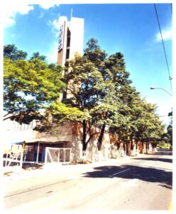 Vista de rua com edifício de torre vertical e letreiro, avenida arborizada em primeiro plano — P0131 — SESC - Piracicaba