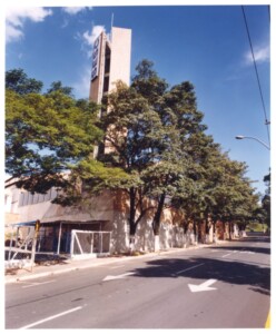 Vista de rua com edifício de torre vertical e letreiro, avenida arborizada em primeiro plano — P0131 — SESC - Piracicaba