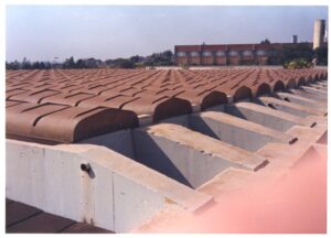 Vista do telhado tipo shed de galpão industrial, mostrando calhas de concreto e cobertura metálica, com edificação ao fundo — P0015 — SESI — Ribeirão Preto