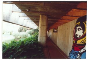 Vista em perspectiva de rampa coberta com piso em degraus baixos, pilar de concreto central, mural artístico na parede lateral e vegetação exuberante sobre o guarda-corpo — P0015 — SESI — Ribeirão Preto