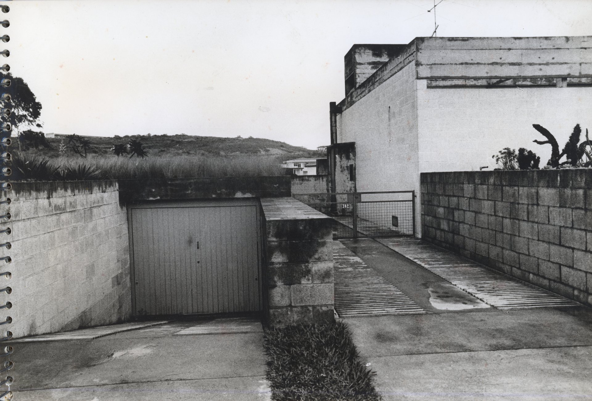 Vista externa de casa mostrando portão de garagem, muro, calçada e número de identificação "162" no portão lateral — P0001 — Residência Teresa Martino — F001-P0001-0000-S03-D90002