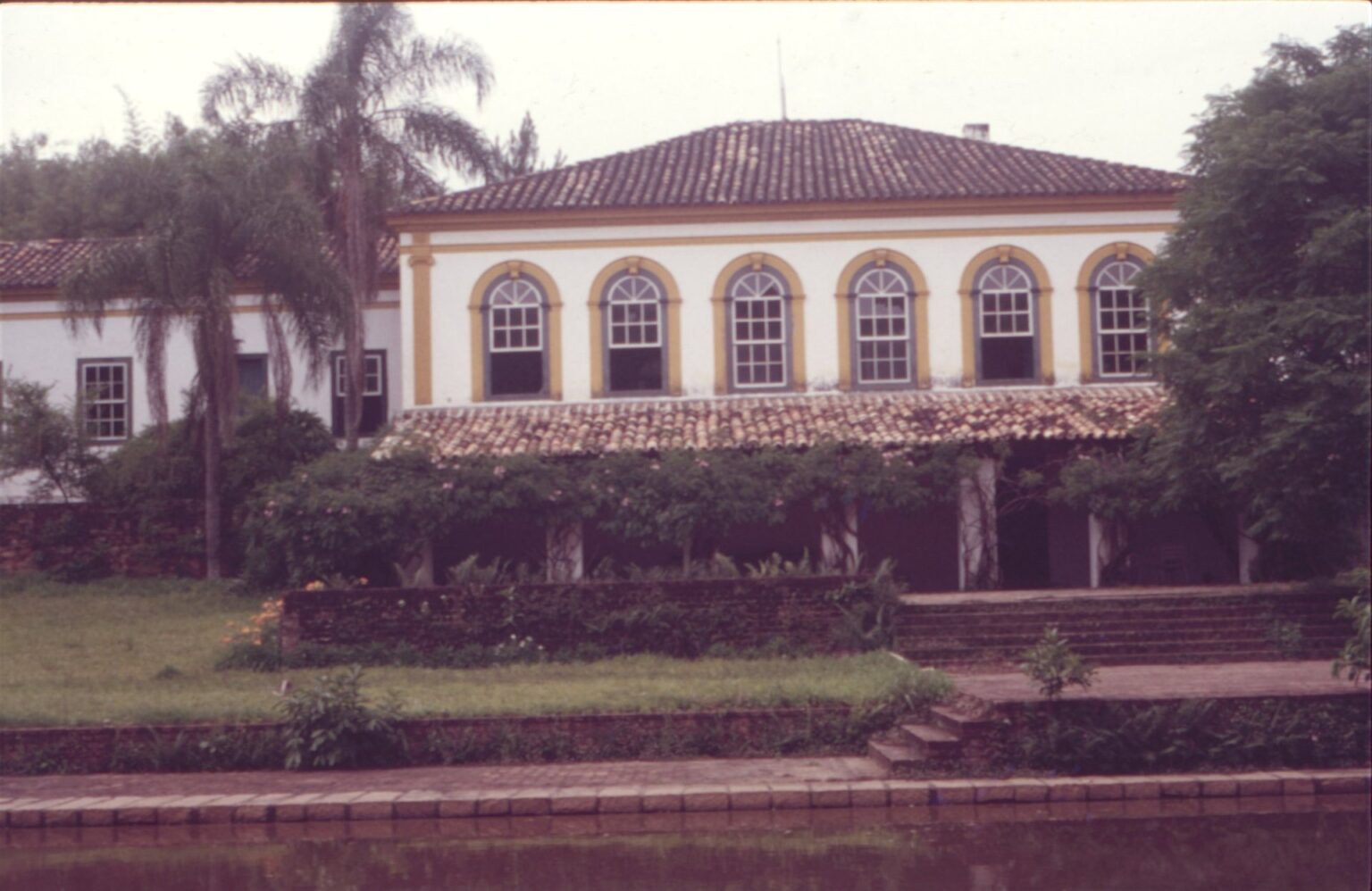Vista externa de casarão colonial com janelas em arco e varanda, junto a um lago — P0024 — Fazenda Fernando Millan — F016-P0024-1981-S01-D00005