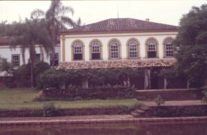 Vista externa de casarão colonial com janelas em arco e varanda, junto a um lago — P0024 — Fazenda Fernando Millan