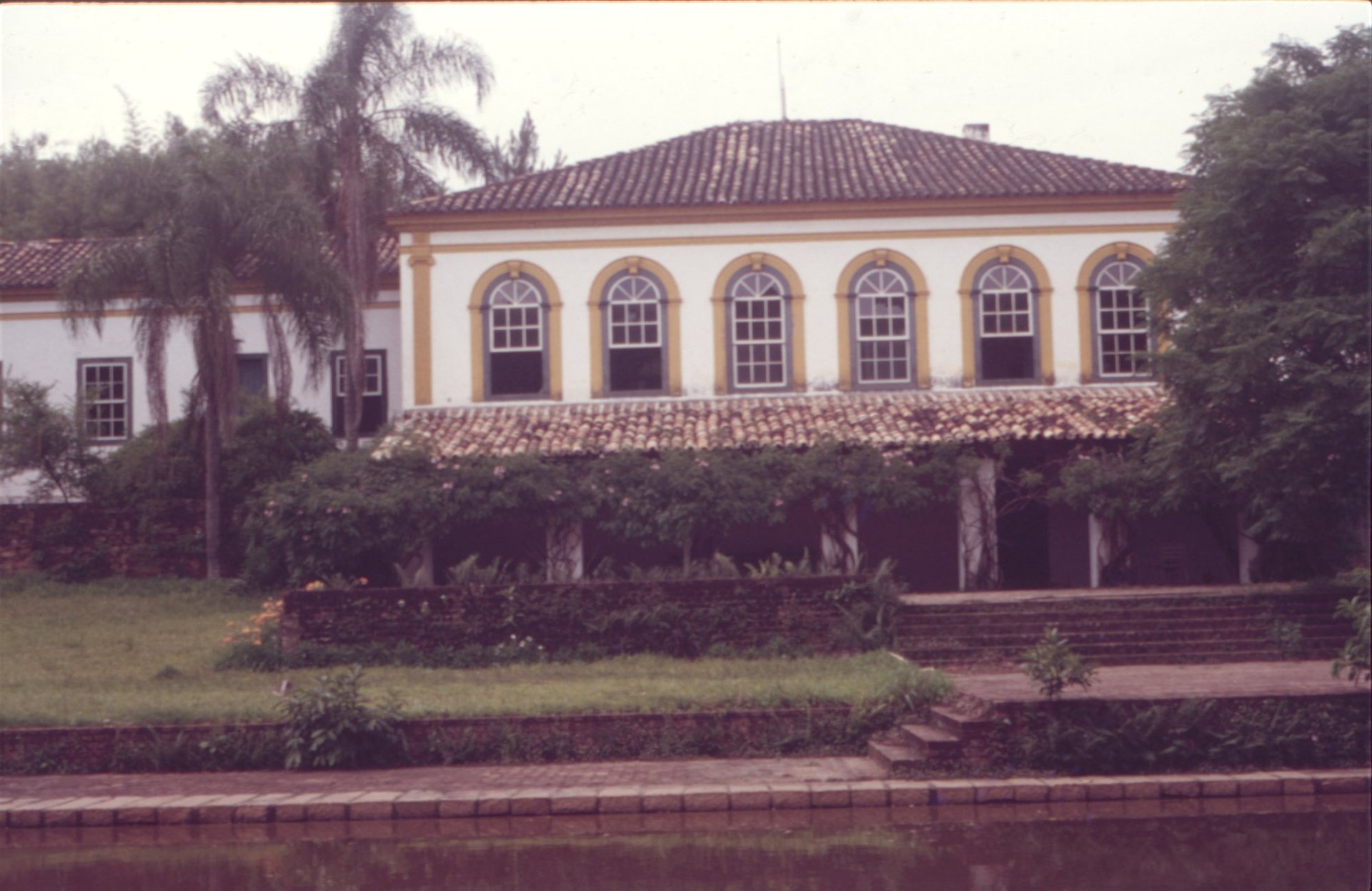 Vista externa de casarão colonial com janelas em arco e varanda, junto a um lago — P0024 — Fazenda Fernando Millan — F016-P0024-1981-S03-D00005