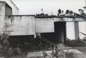 Vista externa de edificação em concreto aparente, mostrando volume em balanço sobre vão de acesso com escada, cobertura ajardinada e jardim tropical em primeiro plano — P0001 — Residência Teresa Martino